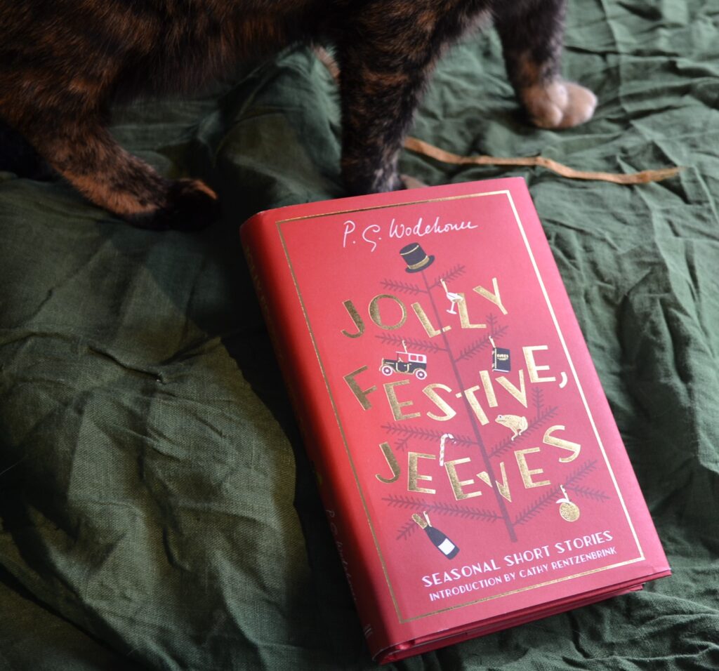 A pair of tortoiseshell paws stand beside a bright red book. Jolly Festive, Jeeves is a collection of seasonal short stories by P G Wodehouse.