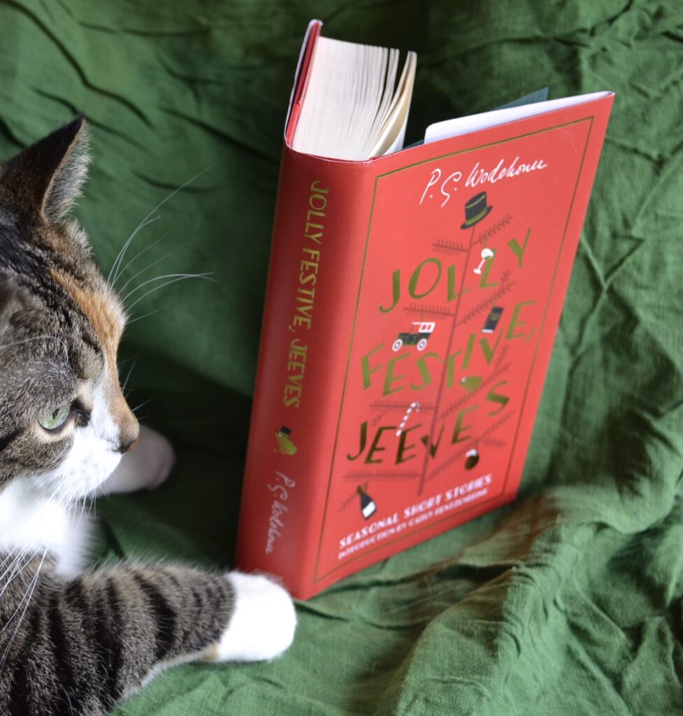 A calico tabby sits on a bright green background with a bright red book. The book's spine has a gold frog wearing a top hat on it.