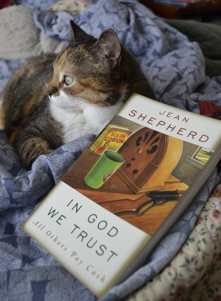 In God We Trust: All Others Pay Cash by Jean Shepherd lies beside a cat and a wooden cardinal.