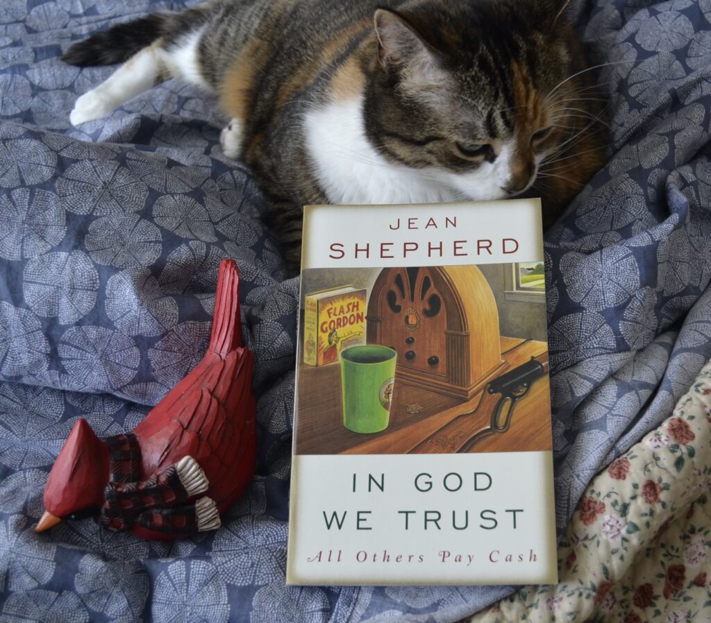 A calico tabby lies beside a wooden cardinal and a book. The book is Jean Shepherd's In God We Trust: All Others Pay Cash.