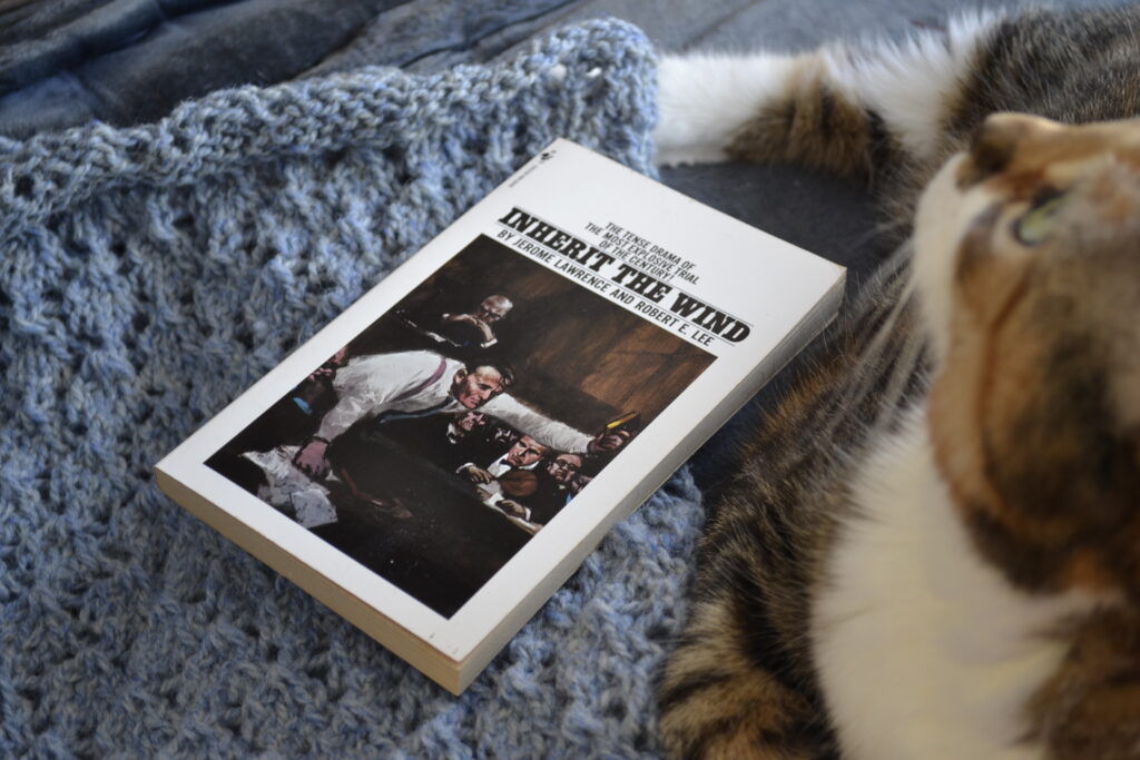 Inherit the Wind by Jerome Lawrence and Robert E Lee is a white book with black text. The cover has a painting of a courtoom with a lawyer holding up a bible.