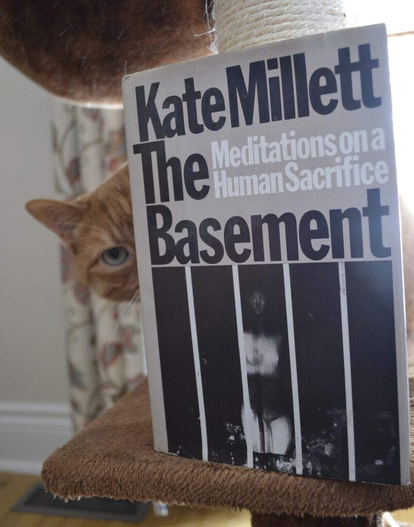 Kate Millett's The Basement: Meditations on a Human Sacrifice is a grey hardcover book. The title and author name are in heavy black font and the picture is a black-and-white photo of a naked woman standing behind bars.