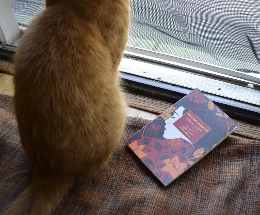 A orange cat sits beside a paperback copy of Written on Water by Eileen Chang.