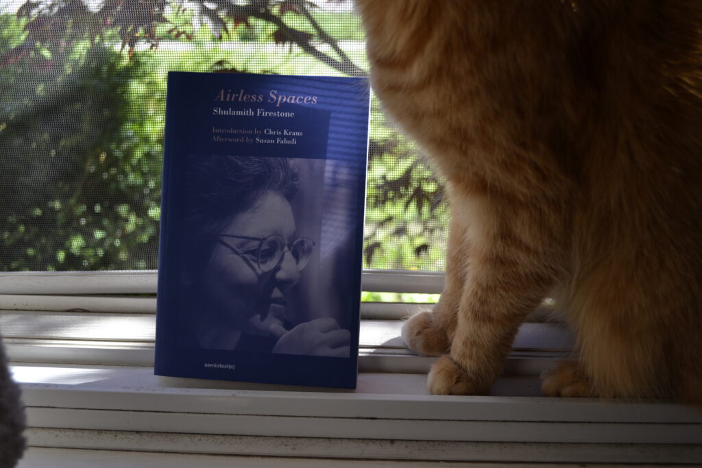 Sulamith Firestone's Airless Spaces is a blue paperback with a picture of the author on the cover. It sits on a window ledge with an orange tabby.