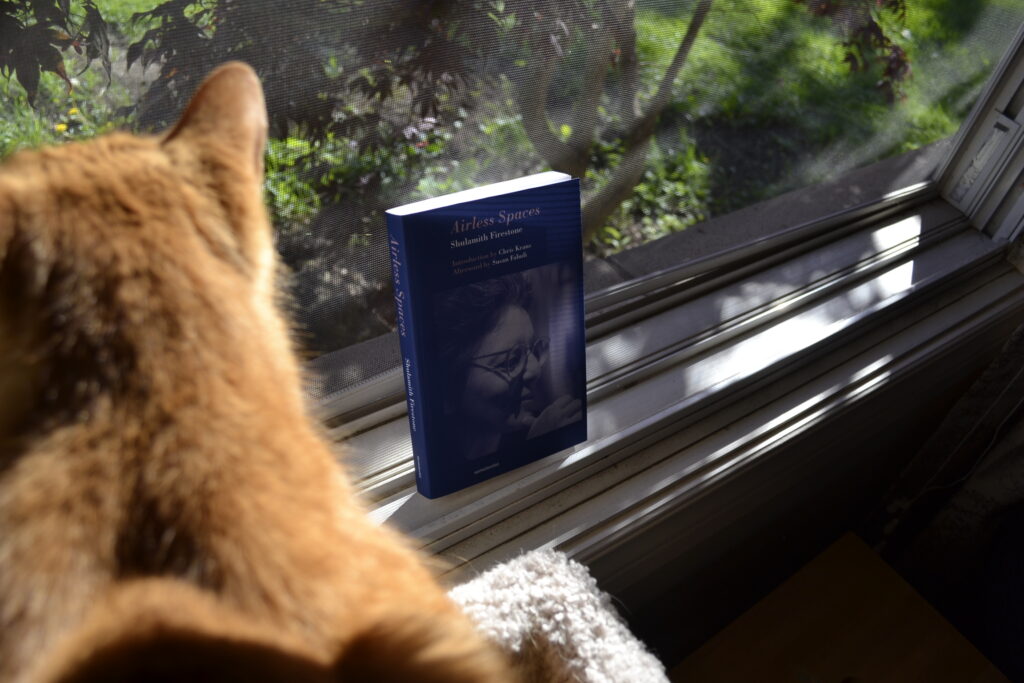 Shulamith Firestone's Airless Spaces is a blue paperback book with a blue-tinted photo of the author, a white woman in her mid-forties wearing round glasses with textured and greying hair that has been brushed back from her face.