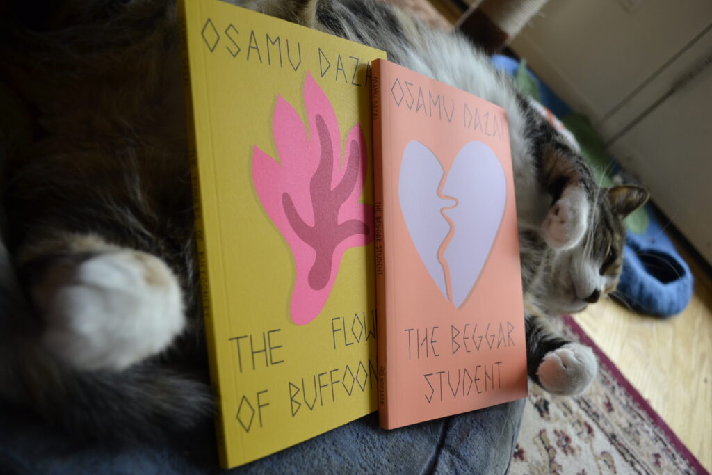 A calico tabby lies on her back. Propped up on her belly are two thin paperbacks, one yellow and one orange.