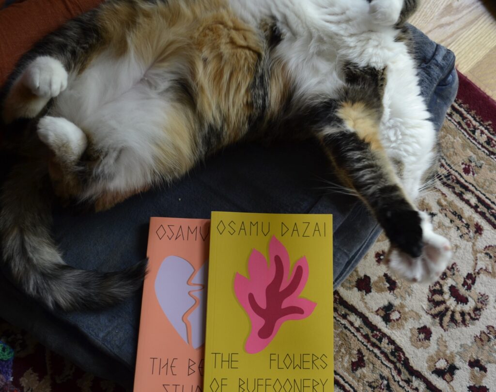 A calico tabby rolls on her back and stretches her paws out, exposing a white and fluffy tummy. Beside her are two books by Osamu Dazai.