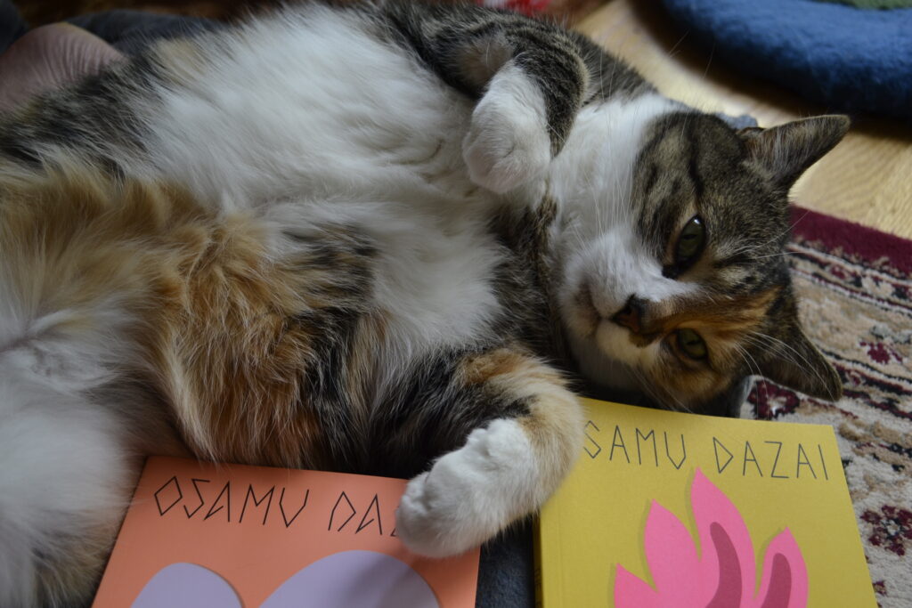 A calico cat lies belly-up with her eyes open but sleepy. Beside her are two thin books by Osamu Dazai.