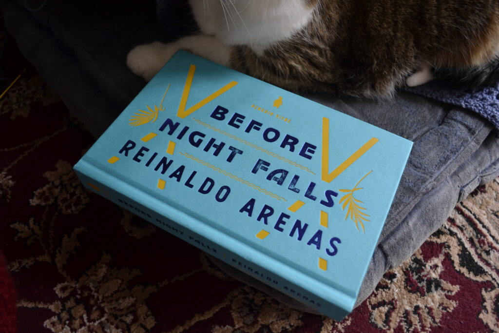 A bright blue hardcover, Before Night Falls by Reinaldo Arenas, nestles against a tabby cat's paws.