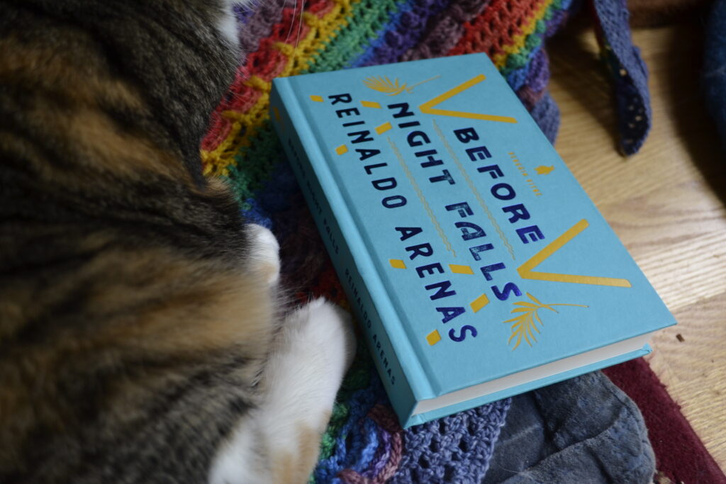 The Penguin Vitae edition of Before Night Falls sits on a rainbow-striped shawl beside a calico cat.