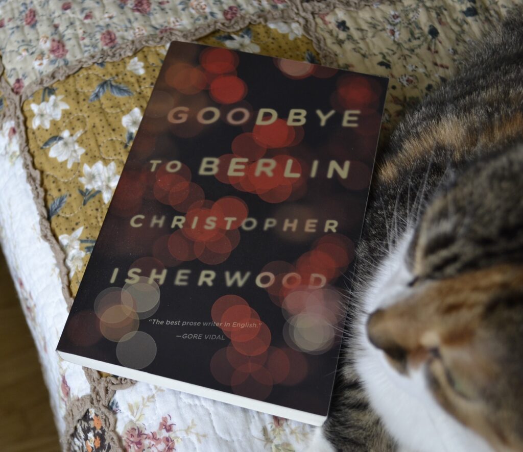 Goodbye to Berlin by Christopher Isherwood lies on a quilt, nestled beside a calico tabby cat.