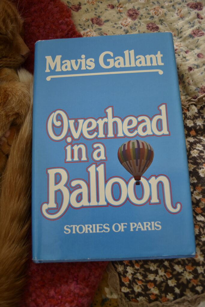 An orange tabby cat curls beside a bright blue book. The book is Overhead in a Balloon: Stories of Paris by Mavis Gallant.