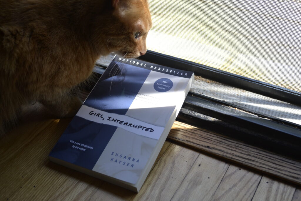 A tabby cat hunches forwards to look at a softcover copy of Girl, Interrupted.