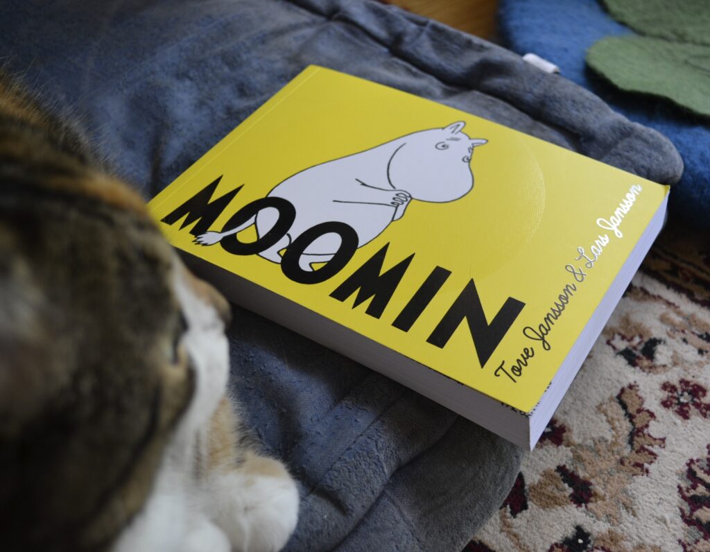 The short and wide graphic novel on the blue cushion is Moomin by Tove Jansson and Lars Jasson. It's yellow cover features a white cartoon animal that looks something like a cow or a hippo with a long tail.