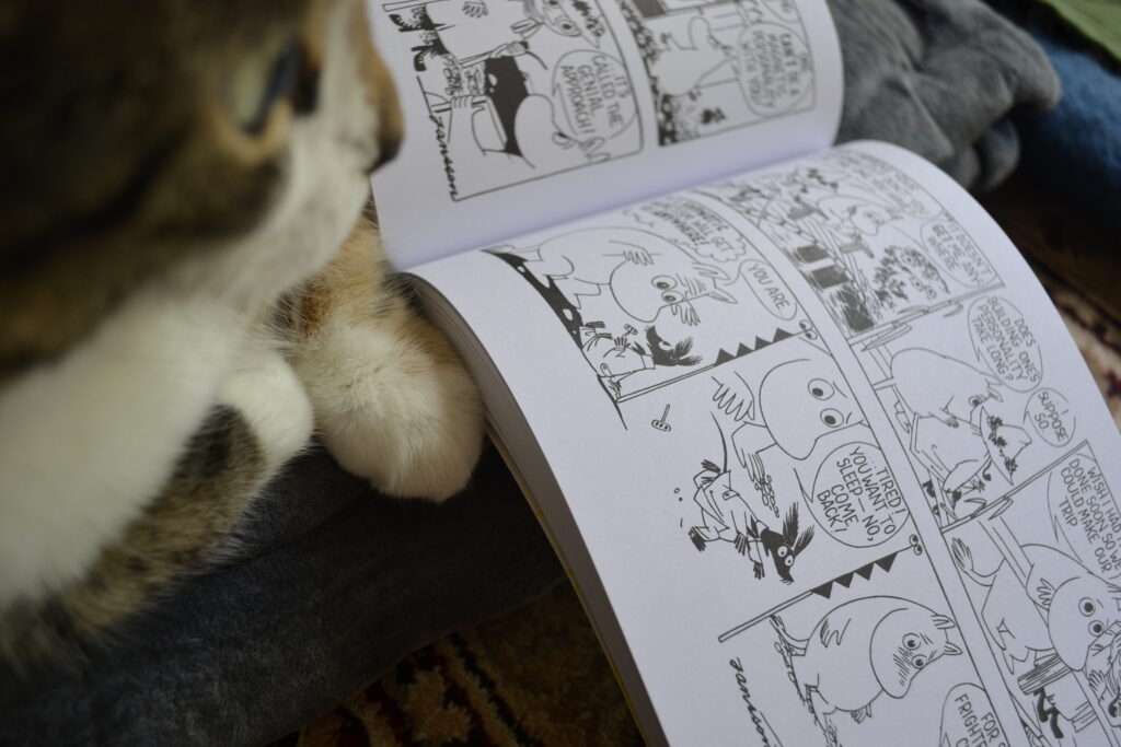 A cat looks through a wide, short graphic novel that is open to show sequential Moomin comics.