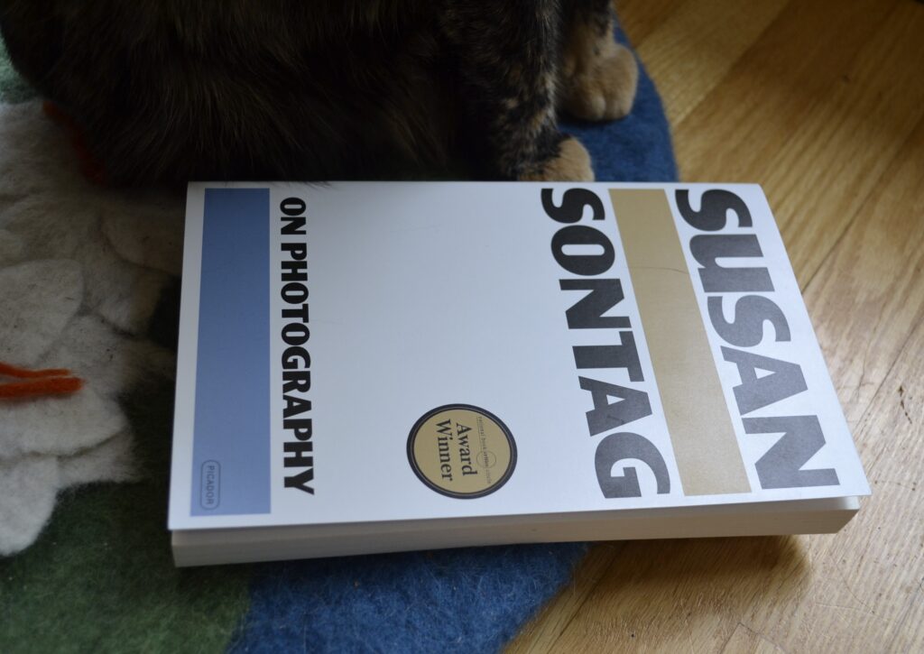 A paperback version of Susan Sontag's On Photography. The cover is plain white with one strip of pale blue and one strip of matte gold. It has an 'Award Winner' sticker on the front.