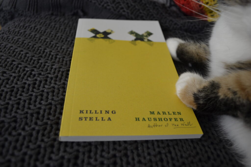 Killing Stella by Marlen Haushofer is a slim yellow paperback. It lies beside the paws of a calico tabby.