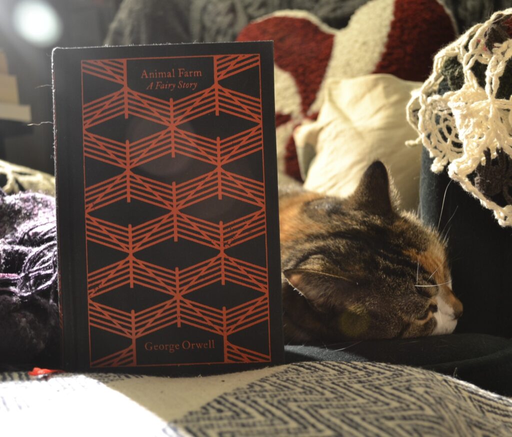 A calico tabby lies flat on a green armchair. In front of her stands a black book, Animal Farm by George Orwell.