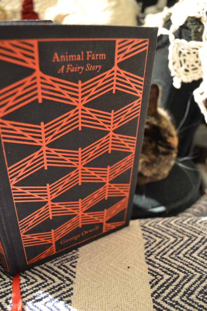 Animal Farm: A Fairy Story by George Orwell is a black and red hardcover. It sits on a green armchair with a calico cat.