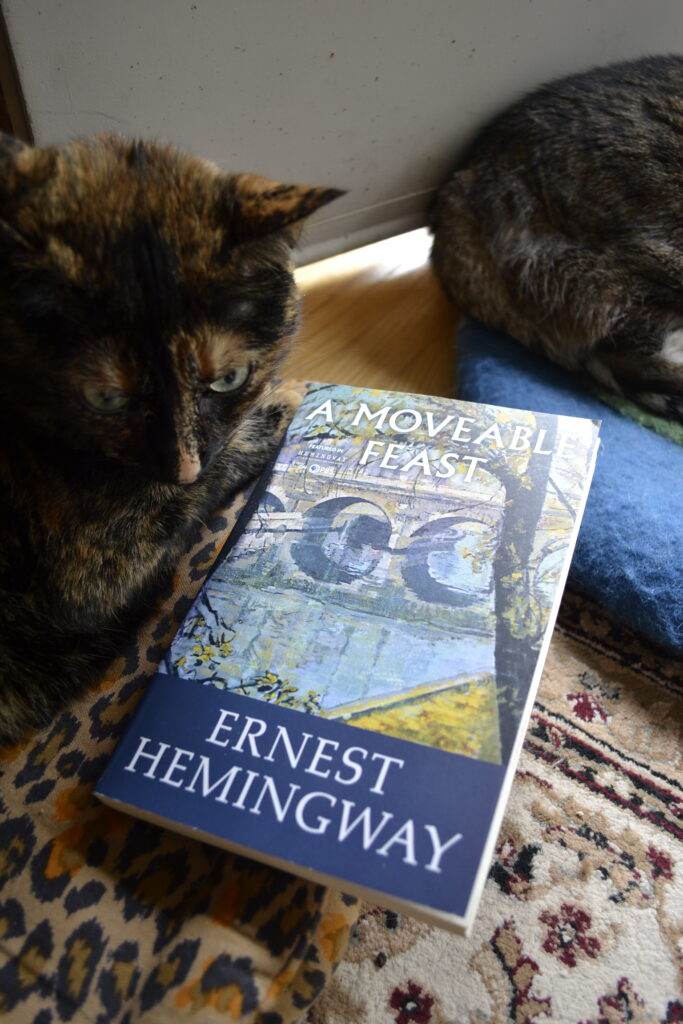 A Moveable Feast by Ernest Hemingway sits between two cats.