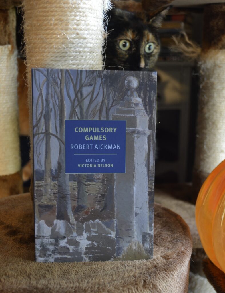 A tortie peeks over the top of a book with a painting of a graveyard on the cover.