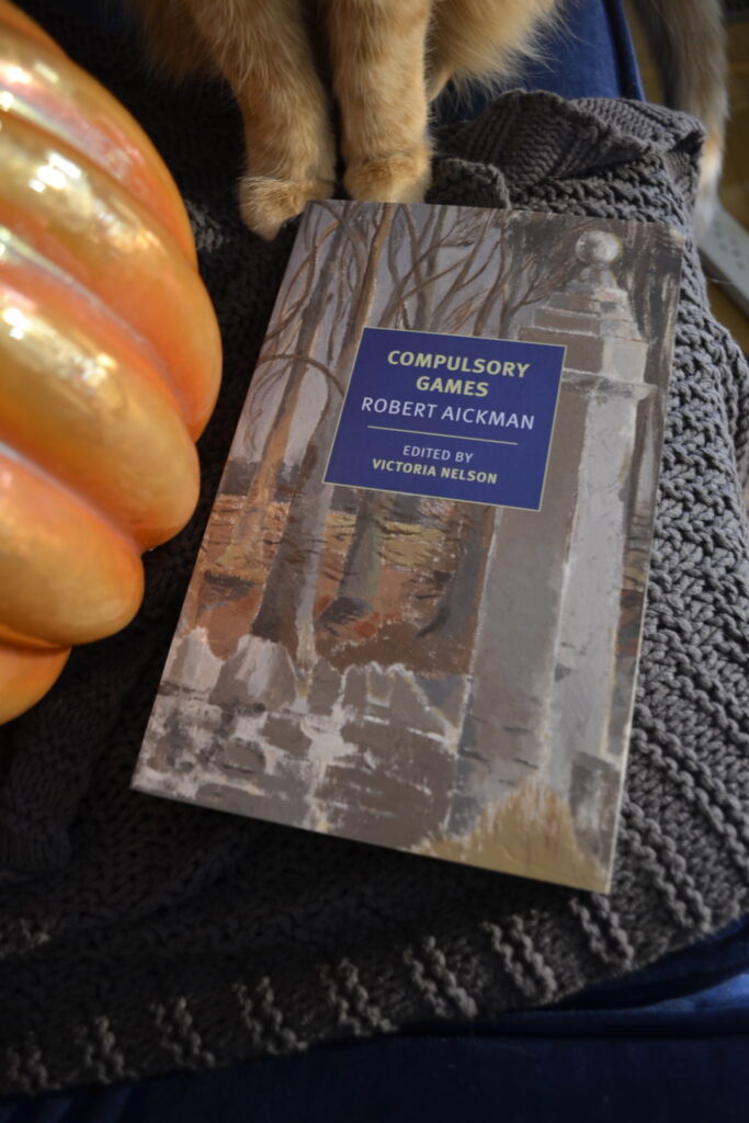 Compulsory Games by Robert Aickman sits beside a glass pumpkin and the paws of an orange cat.