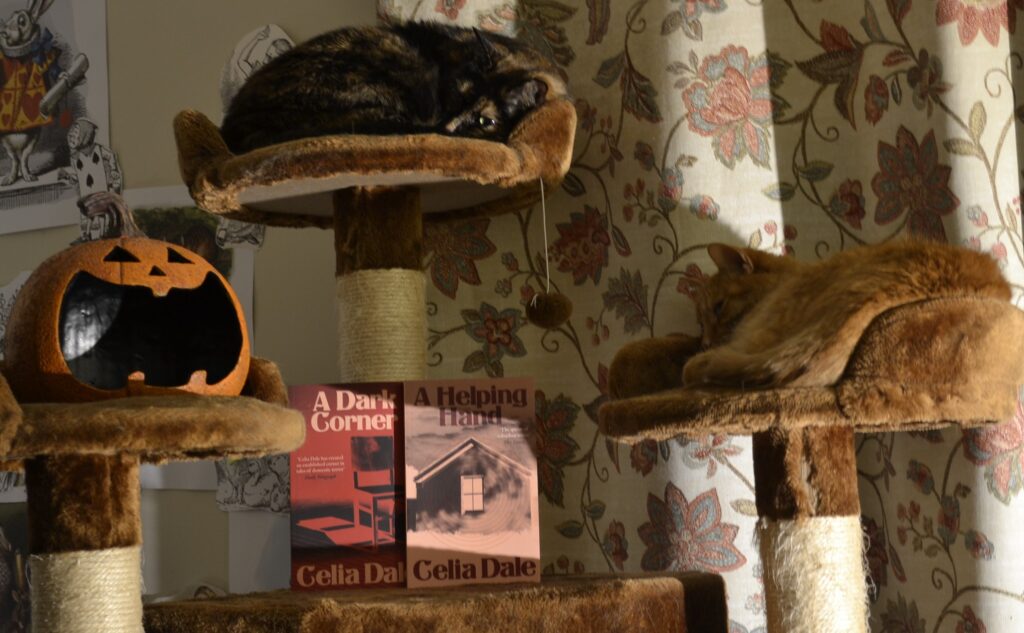 Two books by Celia Dale sit on a cat tree with two cats and a jack-o-lantern decoration that has a huge smile.