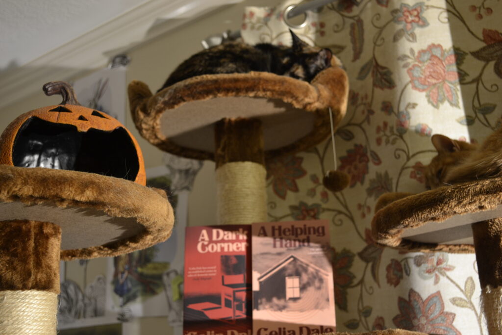 Two complementing books in pink and dark red sit on a cat tree with two cats and a smiling jack-o-lantern decoration.