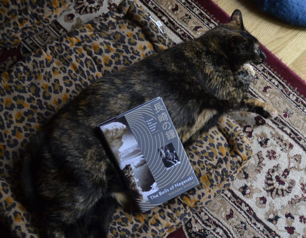 A tortoiseshell cat lies out with a paw in her mouth and grey book lying on her side. The book is The Bells of Nagasaki.