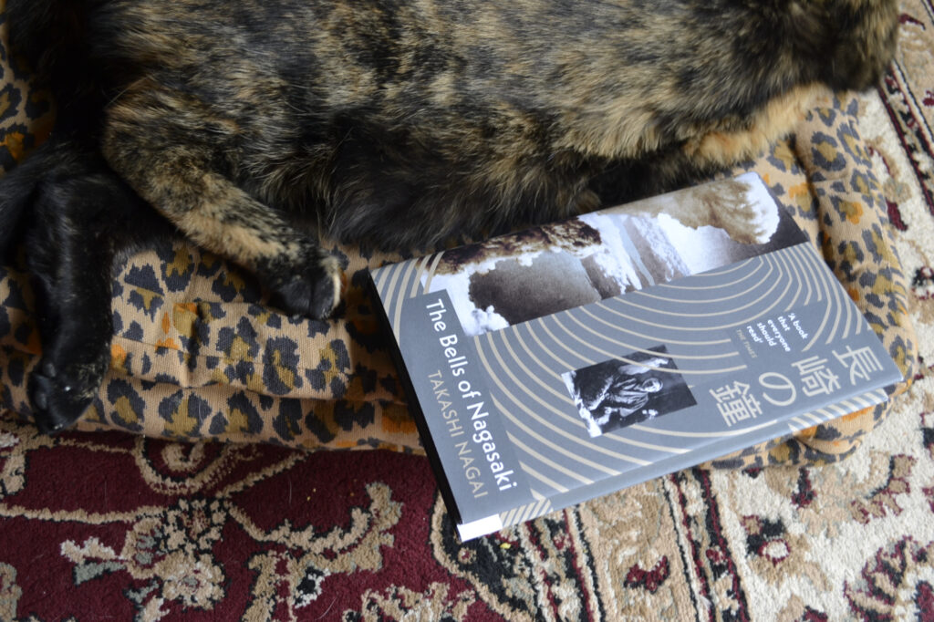 The Bells of Nagasaki is a small, grey hardcover book featuring an image of the atomic bomb explosion and radiating rings.