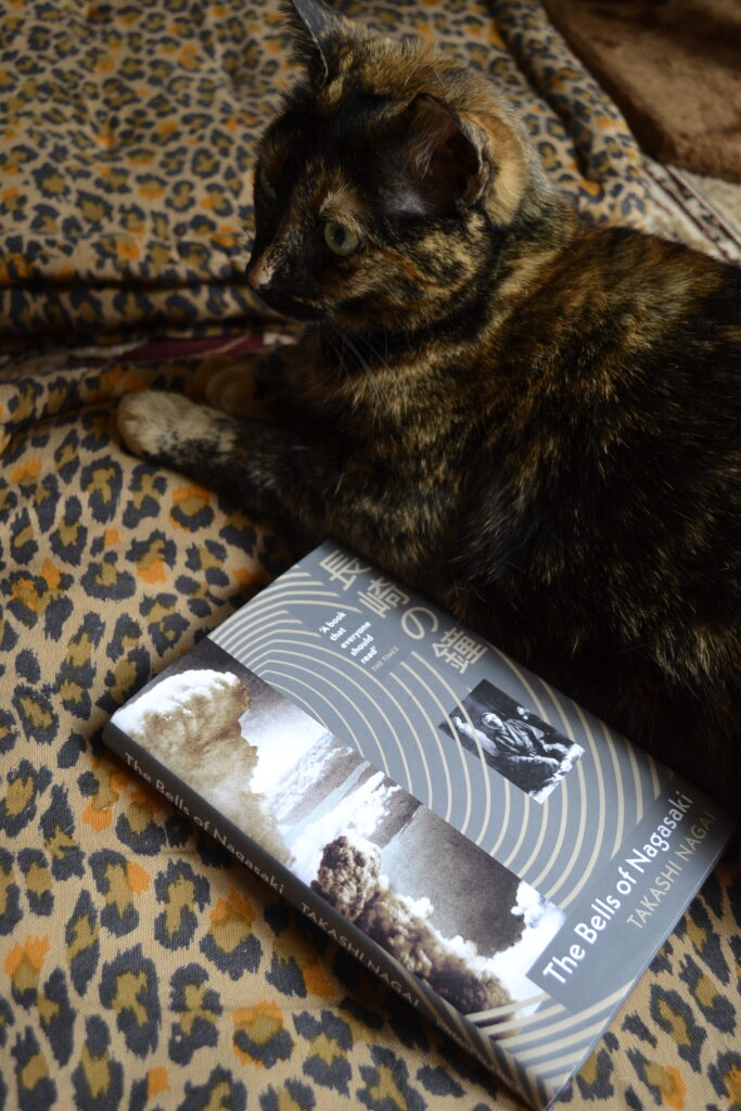 A tortoiseshell cat sits beside a grey book that features Japanese text, radiating rings, and a picture of an atomic explosion.