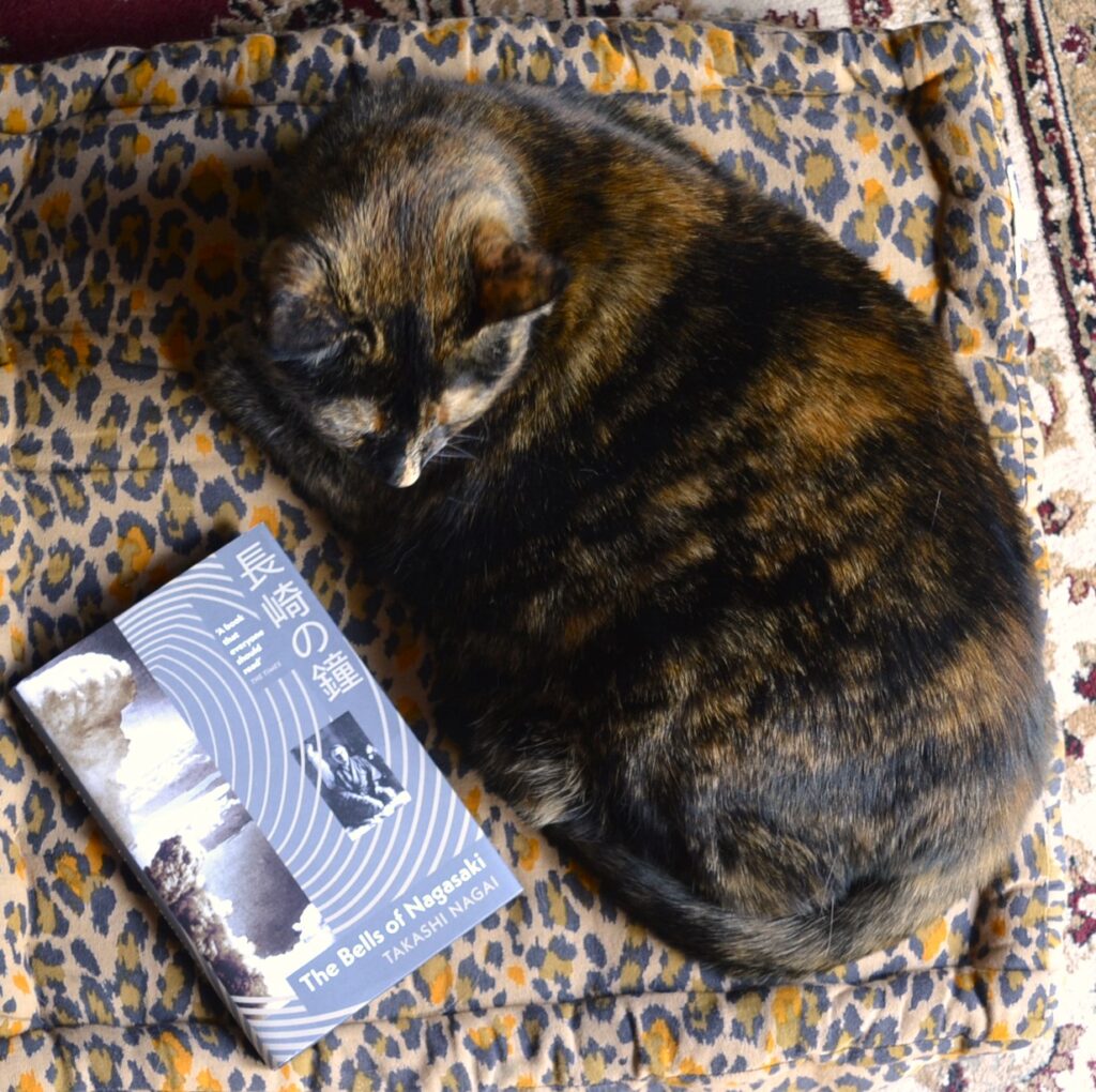 A tortoiseshell cat curls around a copy of The Bells of Nagasaki on a leopard-print cushion.