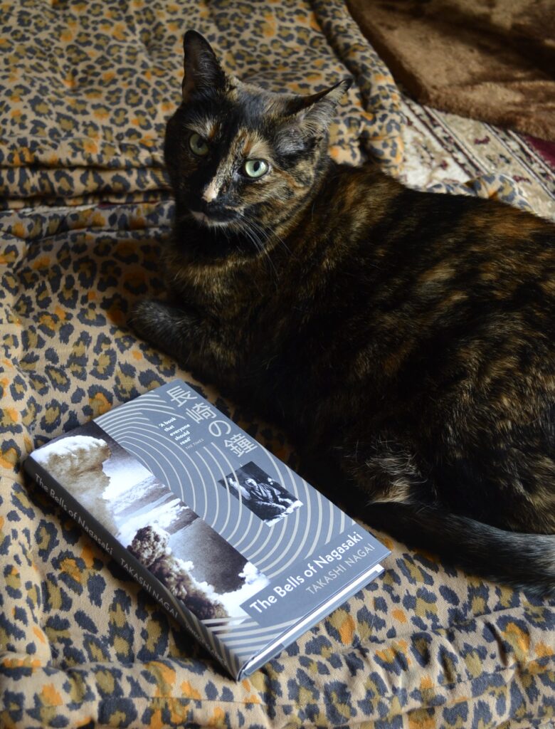 A tortie looks grumpily at you. Beside her is a little, grey, hardcover book with a picture of an atomic explosion on the cover.