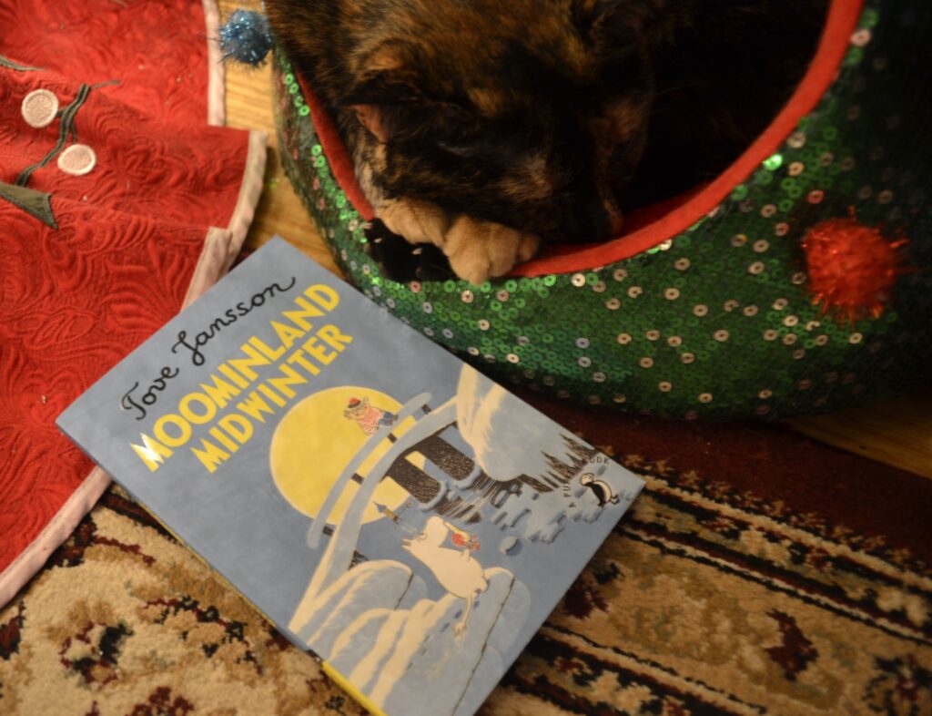 A copy of Moominland Midwinter by Tove Jansson sits beside a tortie that is curled into a Christmas-tree shaped bed.