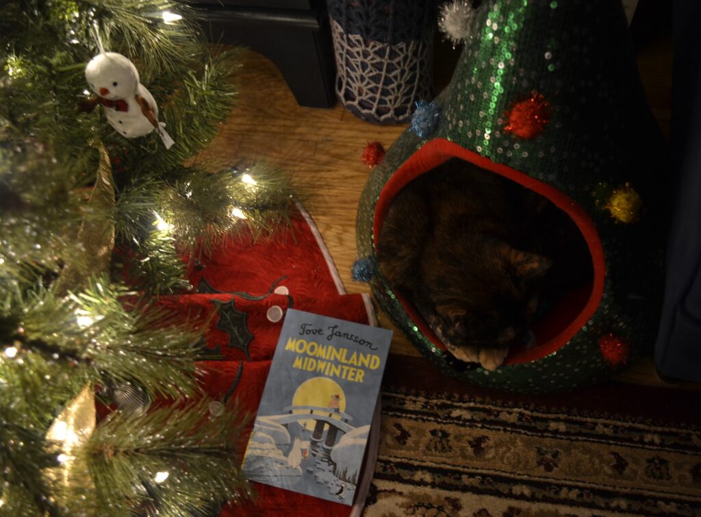 Beneath a lit Christmas tree, a tortoiseshell cat sleeps in a sequined Christmas-tree bed. On the floor beside her is a copy of Moominland Midwinter.