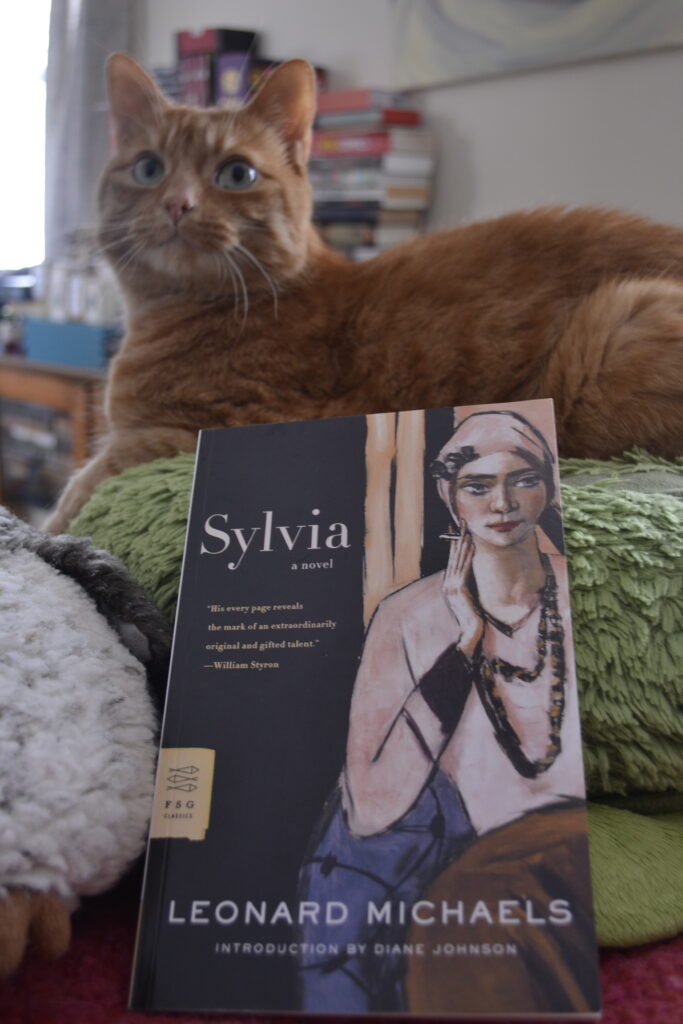 An orange tabby sits primly on a big plushie turtle. In front of her is a book with a woman smoking on the cover. The book is titled Sylvia.