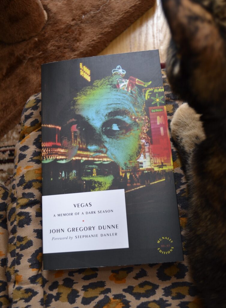 A tortoiseshell cat sits beside a copy of Dunne's Vegas.