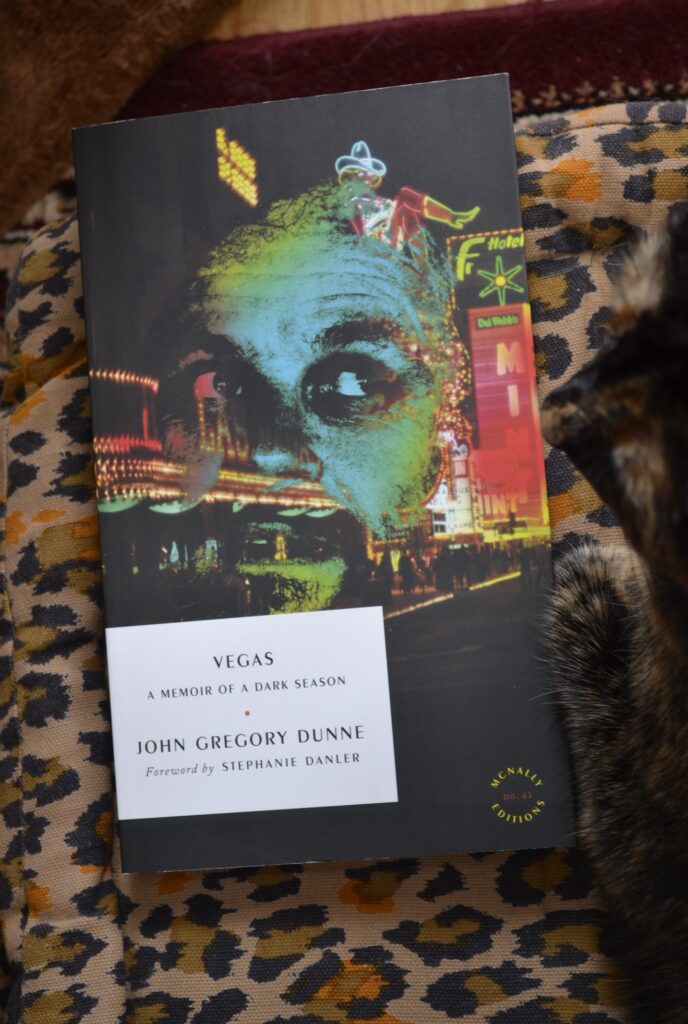 John Gregory Dunne's Vegas is a softcover book featuring a dark picture of neon lights on the Vegas strip overlaid with a neon-blue male face.