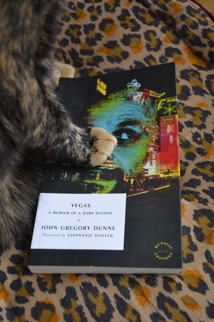 An orange paw presses down on the cover of Vegas by John Gregory Dunne.