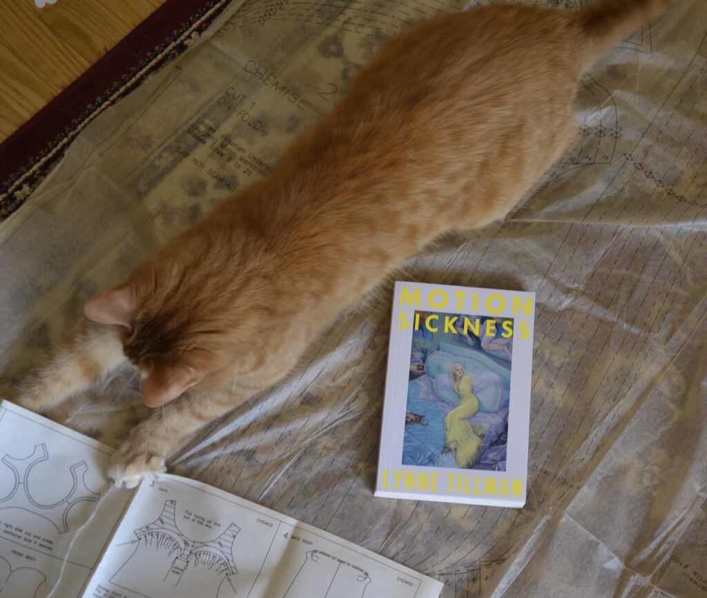 An orange tabby pounces on a sewing pattern. Beside her is a book with a picture of a lounging blonde woman on the cover.