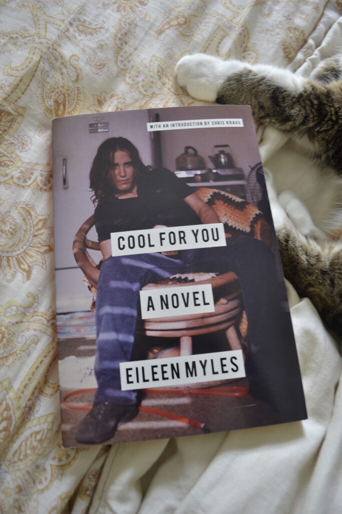 A young woman slouches on a wicker chair on the cover of Eileen Myles' Cool For You. Two white paws are seen pressed against the side of the book.