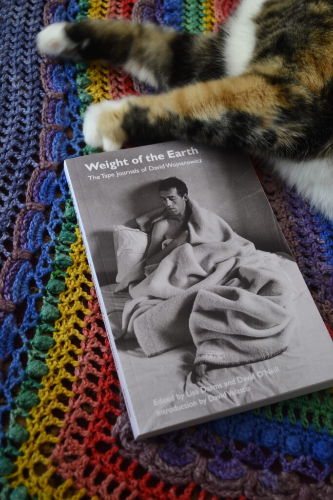 David Wojnarowicz's Weight of the Earth is a black-and-white paperback book lying on a rainbow background.