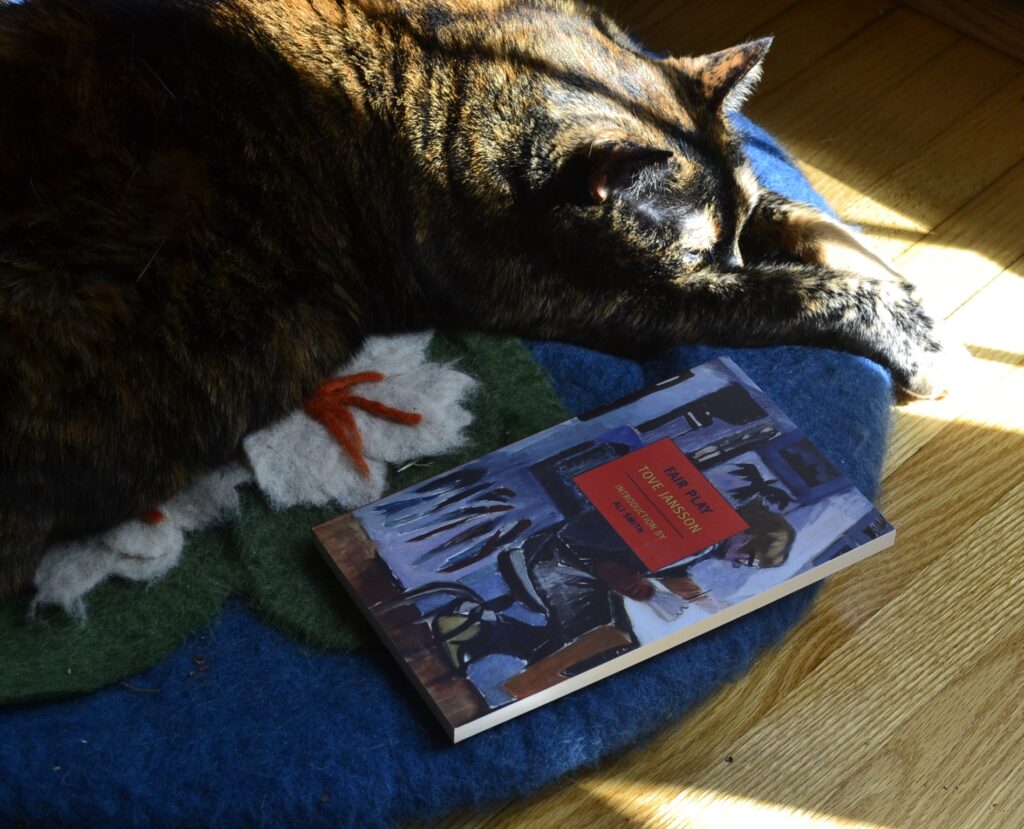 A tortoiseshell cat stretched out her paw and puts her chin between her arms. Beside her, a book with a picture of a person drawing on the cover sits in shadows.