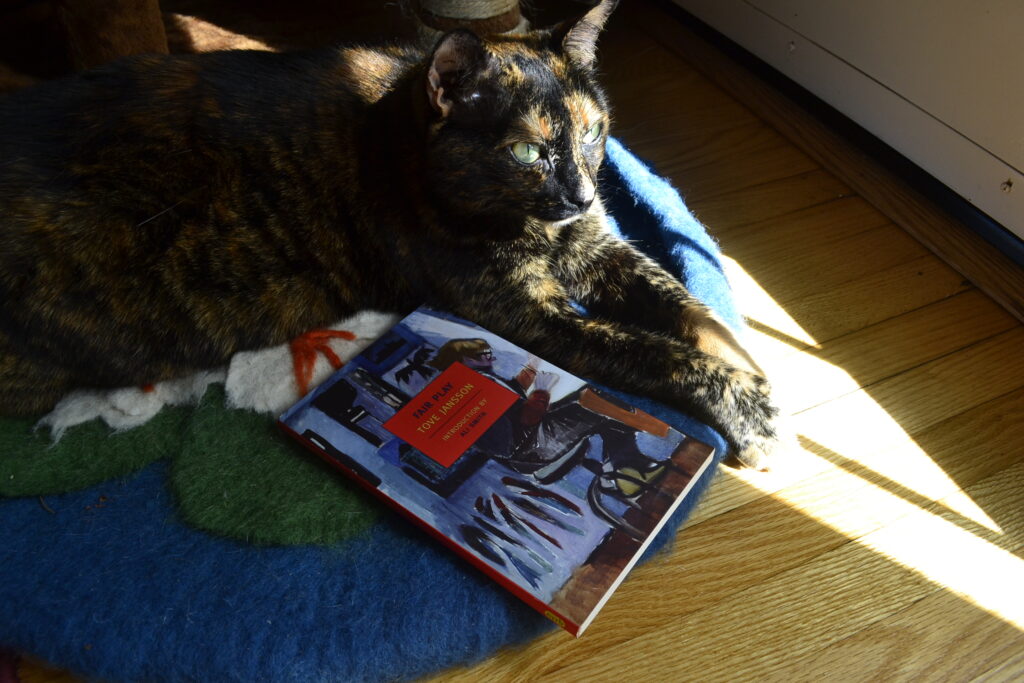 A tortoiseshell cat reached out both her orange paws into the sunlight and holds them together. Beside her is a paperback book by Tove Jansson, Fair Play.