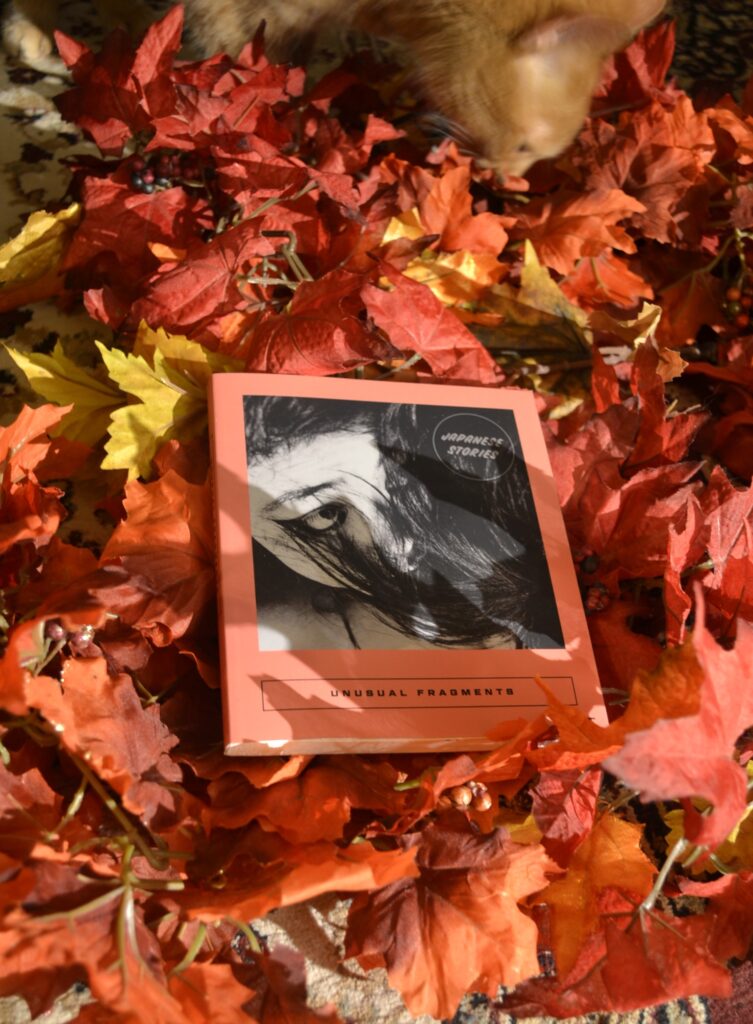 In a pile of red and orange leaves sits a square, coral softcover with a black-and-white picture of an Asian person wearing heavy eyeliner and half-covered by their long, black hair.