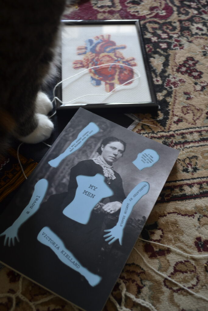 A pair of paws sit between a cross-stitch of an anatomical heart and a paperback copy of My Men.
