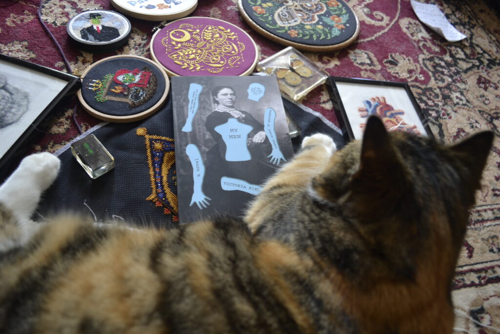 A calico tabby puts her paws on a paperback copy of My Men by Victoria Kielland. Around her are many spooky-themed embroidery projects.