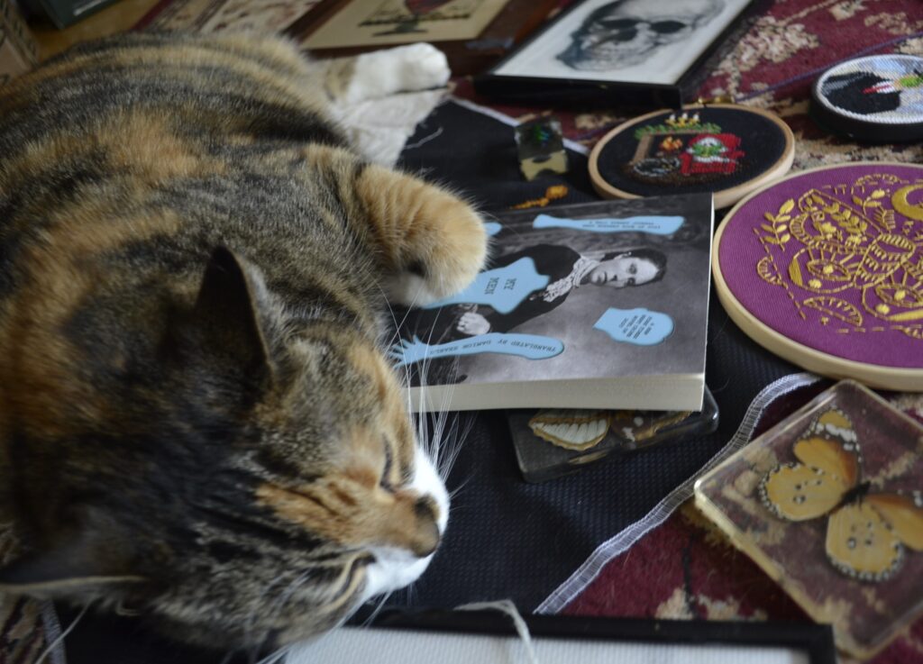 A calico tabby lounges on a pile of embroidery and cross-stitch projects with a book in the middle.