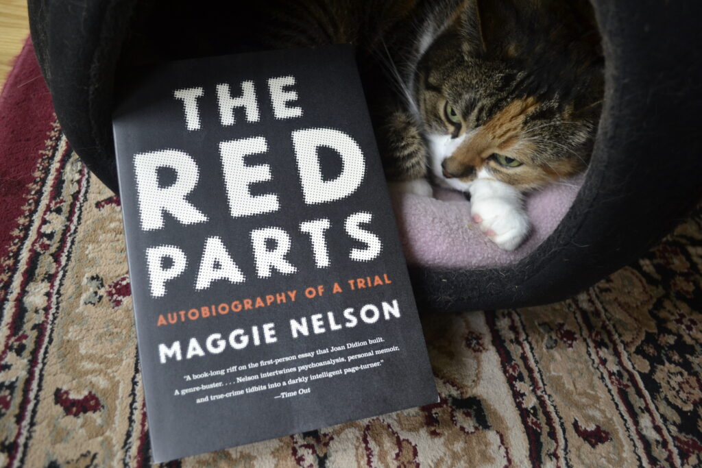 The Red Parts: Autobiography of a Trial is a black softcover book with white font by Maggie Nelson. It lies beside a grumpy calico tabby that's trying to sleep.