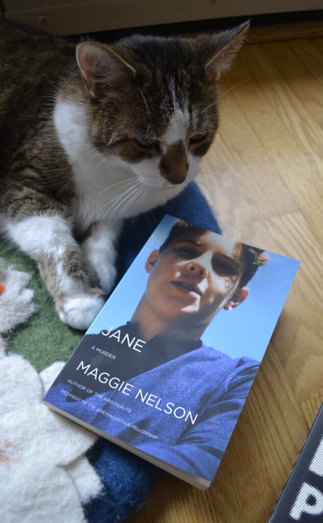 A tabby cat leans over a copy of Jane: A Murder by Maggie Nelson.
